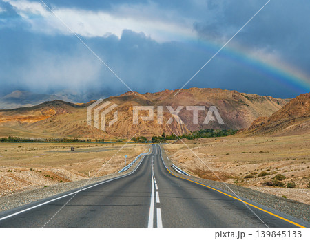 An empty asphalt road stretches through dry Altai mountains. A colorful rainbow brightens the dark, cloudy sky after rain An empty asphalt road stretches through dry Altai mountains. A colorful rainbow brightens the dark, cloudy sky after rain 139845133