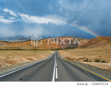 An empty asphalt road stretches through dry Altai mountains. A colorful rainbow brightens the dark, cloudy sky after rain An empty asphalt road stretches through dry Altai mountains. A colorful rainbow brightens the dark, cloudy sky after rain 139845134