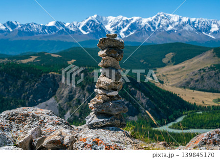 A cairn of rough rocks stands on a high mountain point, showcasing the expansive Altai landscape with its distant snowy peaks and a river 139845170