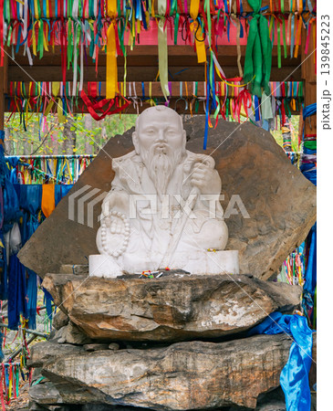 Buryat Shamanic shrine features white stone elder statue holding beads surrounded by vibrant prayer ribbons 139845223