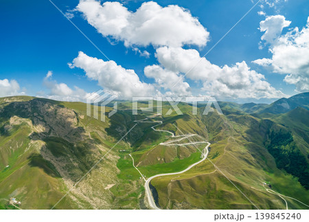 Aerial view showcasing a serpentine road winding through vibrant green mountain valleys between majestic Caucasus mountains Aerial view showcasing a serpentine road winding through vibrant green mountain valleys between majestic Caucasus mountains 139845240