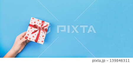 Woman hands give wrapped valentine or other holiday handmade present in paper with red ribbon. Present box, red heart decoration of gift on blue table, top view with copy space for you design Woman hands give wrapped valentine or other holiday handmade present in paper with red ribbon. Present box, red heart decoration of gift on blue table, top view with copy space for you design 139846311