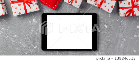 Digital tablet blank screen with gift box and hearts decor on gray cement table. Top view. Valentines Day concept background Digital tablet blank screen with gift box and hearts decor on gray cement table. Top view. Valentines Day concept background 139846315