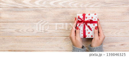 Woman hands give wrapped valentine or other holiday handmade present in paper with red ribbon. Present box, red heart decoration of gift on wooden table, top view with copy space for you design 139846325
