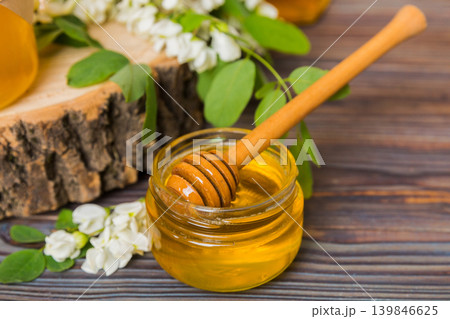 Sweet honey jar surrounded spring acacia blossoms. Honey flows from a spoon in a jar. jars of clear fresh acacia honey on wooden background Sweet honey jar surrounded spring acacia blossoms. Honey flows from a spoon in a jar. jars of clear fresh acacia honey on wooden background 139846625