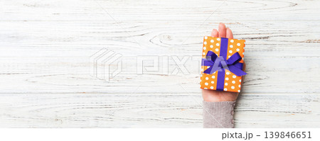 Woman hands give wrapped christmas or other holiday handmade present in colored paper . Present box, decoration of gift on white rustic wooden table, top view with copy space 139846651