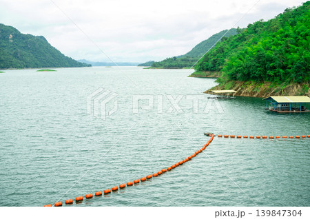 Orange safety buoy line stretching across calm reservoir lake. Green mountains. Water boundary and navigation safety measures. Mountain lake reservoir with floating structures and dense green forest. Orange safety buoy line stretching across calm reservoir lake. Green mountains. Water boundary and navigation safety measures. Mountain lake reservoir with floating structures and dense green forest. 139847304