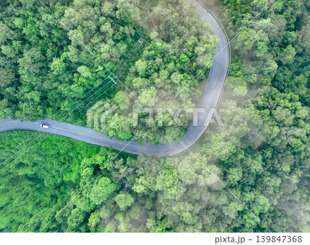 Aerial top view of electric vehicle driving on a winding highway road in green forest with fog. Sustainable transportation, zero emissions, and eco-friendly mobility for reducing carbon footprints. 139847368