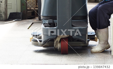 Industrial floor scrubber machine in facility with worker wearing white rubber boots resting after cleaning process. Janitorial service maintenance hygiene and sanitation in workplace environment. 139847432