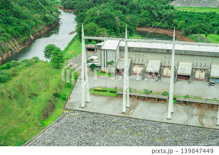 Hydropower plant building with green forest and high-voltage grid infrastructure, with mountain landscape in background. Hydroelectric power station at dam site. Renewable energy. Power generation. Hydropower plant building with green forest and high-voltage grid infrastructure, with mountain landscape in background. Hydroelectric power station at dam site. Renewable energy. Power generation. 139847449