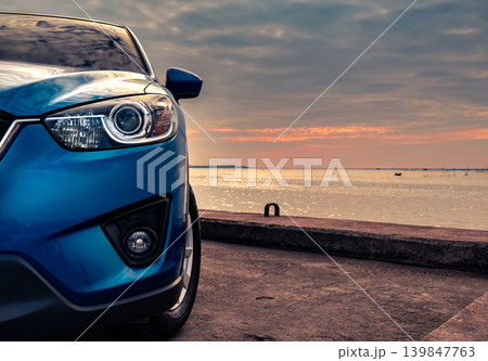 Blue compact SUV car with sport and modern design parked on concrete road by the sea at sunset in the evening. Hybrid and electric car technology concept. Car parking space. Automotive industry. Blue compact SUV car with sport and modern design parked on concrete road by the sea at sunset in the evening. Hybrid and electric car technology concept. Car parking space. Automotive industry. 139847763