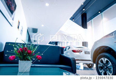 Fake red flowers in vase on table in modern car showroom. Car dealership interior building. Artificial flower on blurred background of new white luxury car parked in showroom. Automotive industry. 139847830