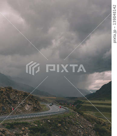 Cars drive along a curving mountain road with a metal guardrail. Dark, heavy clouds fill the sky, casting a moody light over the rugged Altai landscape 139848142