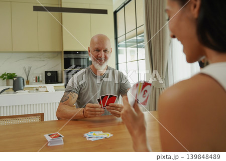 A man and a woman play cards at a wooden table in a sunlit modern living room A man and a woman play cards at a wooden table in a sunlit modern living room 139848489