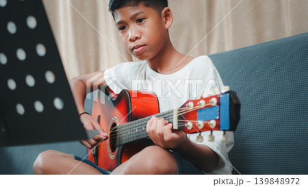 Concentrated Asian boy practicing acoustic guitar while looking at music sheet stand at home. Concept of music education, hobby, childhood development and home leisure activities. 139848972