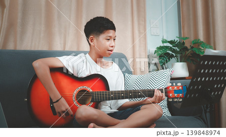 Asian boy practicing acoustic guitar on sofa at home, looking at music sheet stand. Child learning musical instrument, hobby and lifestyle concept for education and talent development. Asian boy practicing acoustic guitar on sofa at home, looking at music sheet stand. Child learning musical instrument, hobby and lifestyle concept for education and talent development. 139848974