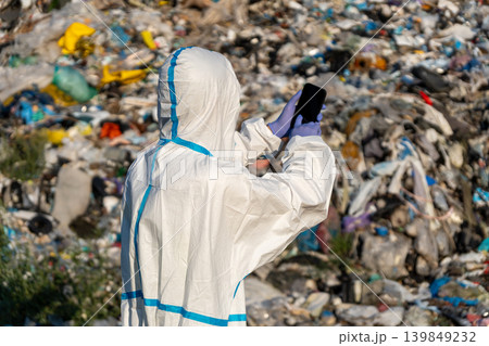 Ecologist wearing protective gear observes and records data at a municipal landfill filled with waste and debris during daylight hours 139849232