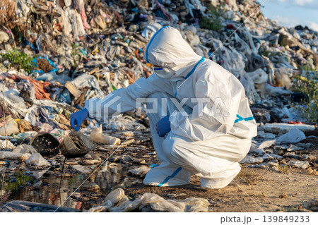 Environmentalist in protective gear checks waste and pollution levels at a landfill site while collecting samples from water 139849233
