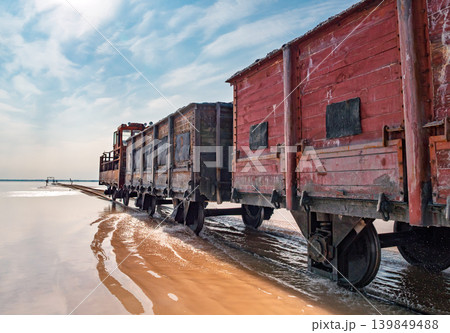 Old, rusty locomotive and wooden freight wagons travel along tracks through the shallow, pink water of Bursol Lake Old, rusty locomotive and wooden freight wagons travel along tracks through the shallow, pink water of Bursol Lake 139849488
