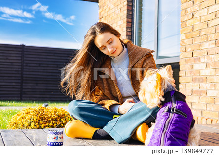 A happy young woman sitting cross-legged outdoors with her small dog on a sunny day, cozy autumn outfit and calm lifestyle scene, pet human friendship A happy young woman sitting cross-legged outdoors with her small dog on a sunny day, cozy autumn outfit and calm lifestyle scene, pet human friendship 139849777