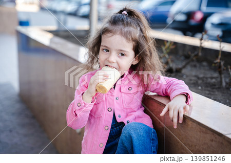 Little Caucasian girl eating ice cream outdoors while wearing a pink jacket and sitting in an urban residential area. Childhood lifestyle Little Caucasian girl eating ice cream outdoors while wearing a pink jacket and sitting in an urban residential area. Childhood lifestyle 139851246