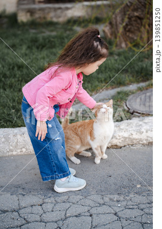Little Caucasian girl petting a ginger cat on a residential street while showing care and affection for domestic animals outdoors. Childhood lifestyle 139851250