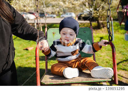 Curious toddler boy sitting on a playground swing while holding an adult hand outdoors in sunny daylight. Childhood lifestyle Curious toddler boy sitting on a playground swing while holding an adult hand outdoors in sunny daylight. Childhood lifestyle 139851295