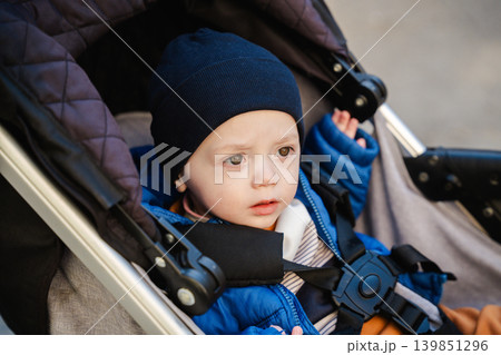 Caucasian toddler boy sitting in a stroller outdoors while wearing a blue jacket and black hat with safety straps fastened securely. Family lifestyle Caucasian toddler boy sitting in a stroller outdoors while wearing a blue jacket and black hat with safety straps fastened securely. Family lifestyle 139851296