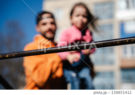 Father supporting his little Caucasian daughter near playground bars outdoors with a blurred background in a sunny urban area. Family lifestyle Father supporting his little Caucasian daughter near playground bars outdoors with a blurred background in a sunny urban area. Family lifestyle 139851412