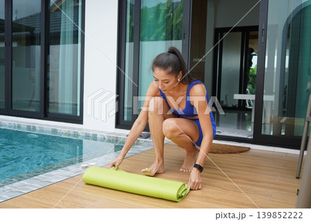 woman rolling yoga mat by pool preparing for morning stretch on deck wearing blue shorts lace woman rolling yoga mat by pool preparing for morning stretch on deck wearing blue shorts lace 139852222