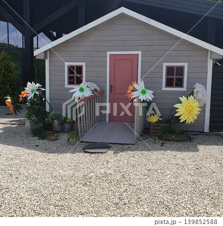A fairy-tale wooden house with a pink door and large artificial flowers in the yard. A cute decorative location A fairy-tale wooden house with a pink door and large artificial flowers in the yard. A cute decorative location 139852588