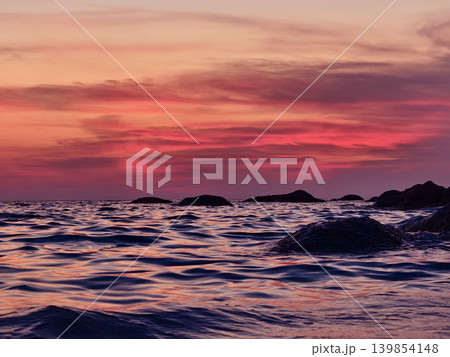 Vibrant sunset over rocky coastal waves, pink and coral sky reflecting on rippling ocean surface. 139854148