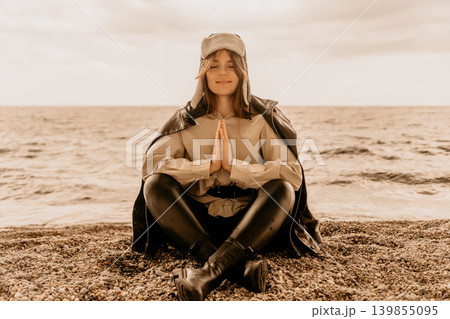 Meditation beach woman sits in lotus pose while practicing mindfulness on a cold pebble shore wearing a warm ushanka hat Meditation beach woman sits in lotus pose while practicing mindfulness on a cold pebble shore wearing a warm ushanka hat 139855095