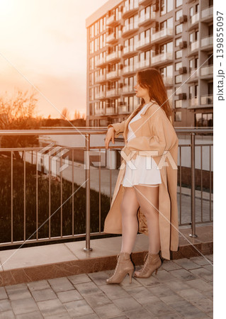 Woman trench coat sunset standing on a balcony of a modern apartment building during the golden hour in the city Woman trench coat sunset standing on a balcony of a modern apartment building during the golden hour in the city 139855097