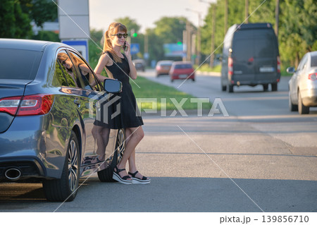 Stylish woman driver standing near her vehicle talking on cellphone on city street in summer Stylish woman driver standing near her vehicle talking on cellphone on city street in summer 139856710