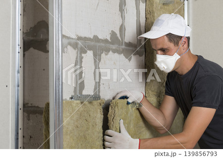 Worker in protective respirator mask insulating rock wool insulation in wooden frame for future house walls for cold barrier. Comfortable warm home, economy, construction and renovation concept Worker in protective respirator mask insulating rock wool insulation in wooden frame for future house walls for cold barrier. Comfortable warm home, economy, construction and renovation concept 139856793