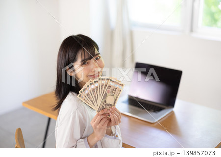 パソコンデスクの前で10枚の1万円札を手に微笑む日本人の若い女性 139857073