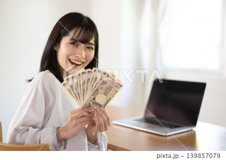 パソコンデスクの前で10枚の1万円札を手に微笑む日本人の若い女性 139857079