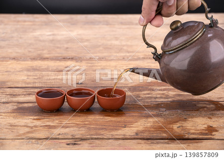 Hand Holding an Antique Brown Teapot Pouring Fresh Hot Tea Into Three Small Traditional Terracotta Clay Cups Arranged in a Row on a Rustic Weathered Wooden Table with Dark Background 139857809