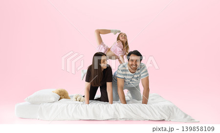 Smiling family playing on white bed with daughter climbing on parents backs. Smiling family playing on white bed with daughter climbing on parents backs. 139858109