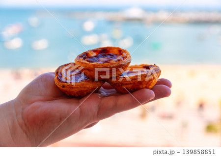 Pastel de nata pastries in hand with blurred Cascais waterfront background Pastel de nata pastries in hand with blurred Cascais waterfront background 139858851