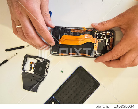 A technician repairs a modern smartphone with exposed internal components, battery, and tools on a white workbench. Concept for electronics repair and mobile device servicing A technician repairs a modern smartphone with exposed internal components, battery, and tools on a white workbench. Concept for electronics repair and mobile device servicing 139859034