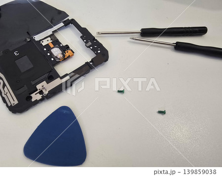 A technician repairs a modern smartphone with exposed internal components, battery, and tools on a white workbench. Concept for electronics repair and mobile device servicing 139859038