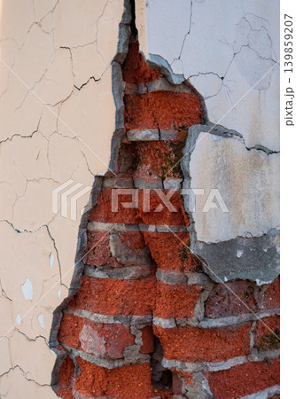 A severely damaged exterior wall shows extensive cracking and peeling of its light-colored plaster. Red bricks and grey mortar are exposed 139859207