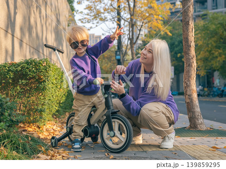 Smiling mother crouches next to her young son on a balance bike on a brick sidewalk. They wear matching purple shirts during a bright autumn afternoon Smiling mother crouches next to her young son on a balance bike on a brick sidewalk. They wear matching purple shirts during a bright autumn afternoon 139859295