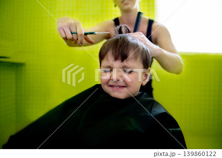 Kid, boy and hairdresser with haircut at salon for grooming, hairstyle and grimace with scissors. Barber, person and child for hair treatment, haircare and hairdressing with comb, happy and grimace 139860122