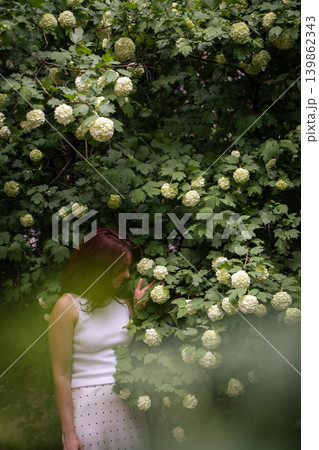 Red-haired woman touches white hydrangea blossom, standing among dense green foliage with soft foreground blur in shaded summer garden. Wellness content, beauty advertising, botanical storytelling Red-haired woman touches white hydrangea blossom, standing among dense green foliage with soft foreground blur in shaded summer garden. Wellness content, beauty advertising, botanical storytelling 139862343