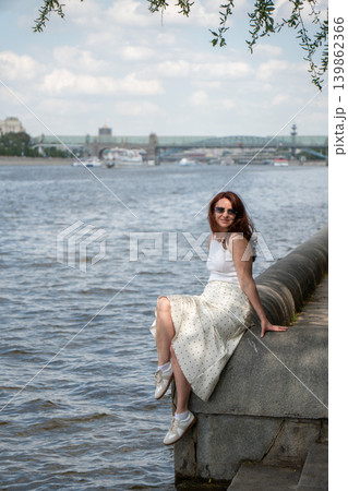 Red-haired woman sits on Gorky Park embankment beside Moscow River with city bridge. Lifestyle marketing, wellness branding, city leisure content, travel advertising, summer recreation Red-haired woman sits on Gorky Park embankment beside Moscow River with city bridge. Lifestyle marketing, wellness branding, city leisure content, travel advertising, summer recreation 139862366