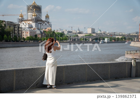 Red-haired woman photographs Cathedral of Christ the Saviour from Moscow River embankment under blue sky with city skyline, water view. Tourism, mobile photography, travel, city landmark, social media 139862371