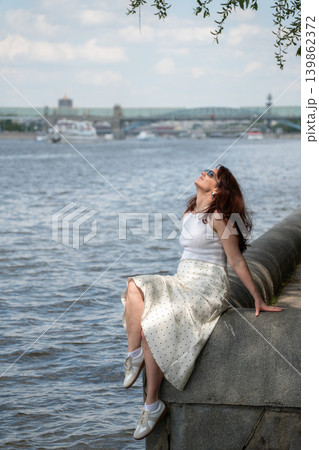 Red-haired woman relaxes on embankment in Gorky Park, Moscow River under warm summer sunlight with urban waterfront. Lifestyle, wellness branding, city leisure, travel advertising, summer recreation Red-haired woman relaxes on embankment in Gorky Park, Moscow River under warm summer sunlight with urban waterfront. Lifestyle, wellness branding, city leisure, travel advertising, summer recreation 139862372
